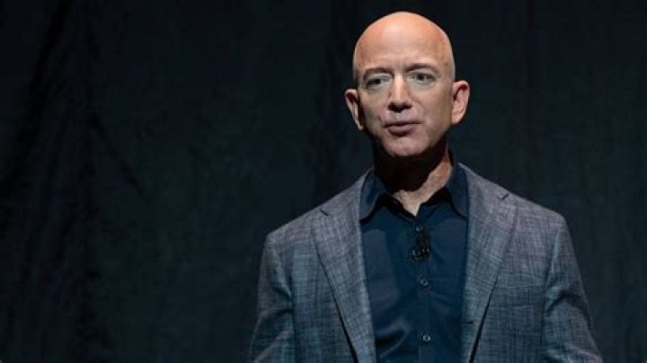 Jeff Bezos in a black and white suite, purple checkered tie, and smiling with his mouth closed. 